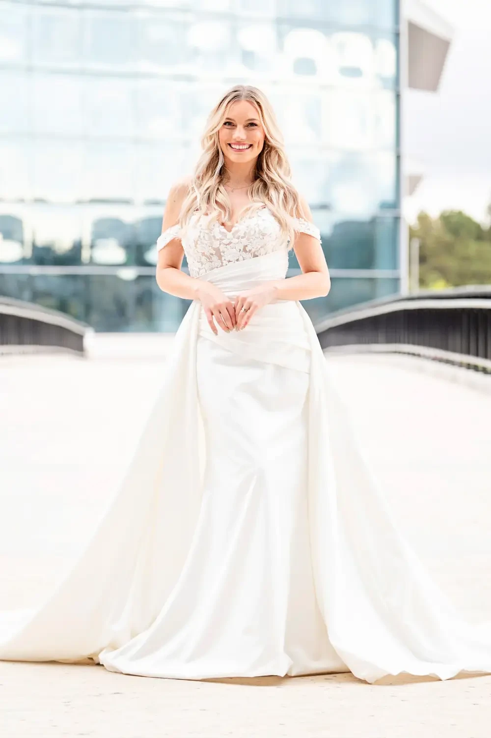 A woman in a wedding dress stands on a bridge with a modern building in the background.