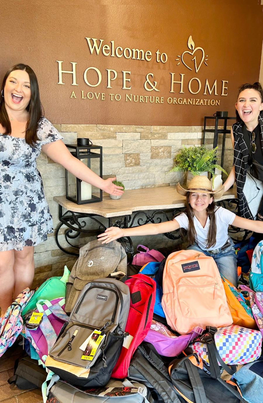 Three women present a large collection of colorful backpacks in front of a wall that reads 
